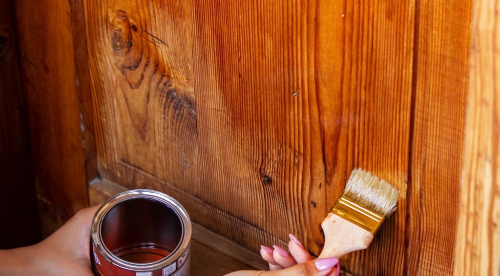 A young woman's hands hold both wood stain and a brush that applies the stain to a section of wood.