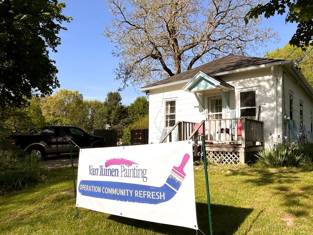 A white home with a yard sign in front advertising that the home is the winner of Van Tuinen Painting's 2023 Operation Community Refresh project.