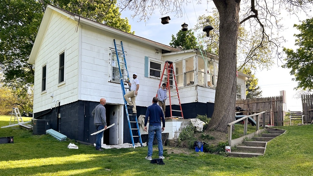 Exterior home professionals work on painting and restoring the exterior of a small, white house.