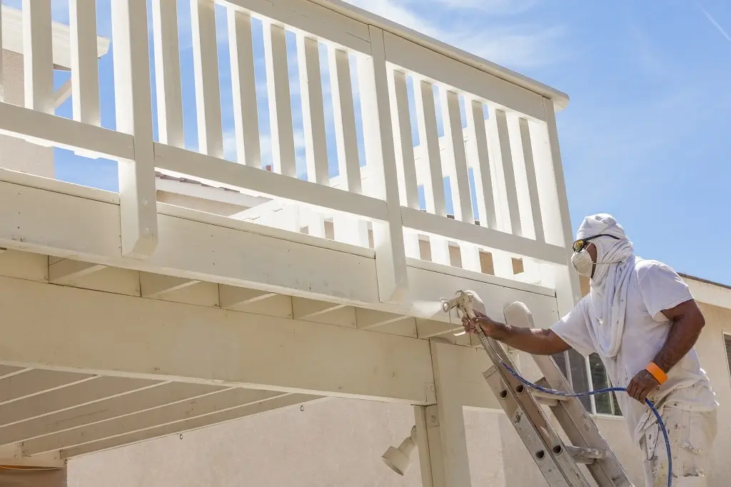 A house painter wearing protection from the sun spray paints a white deck.