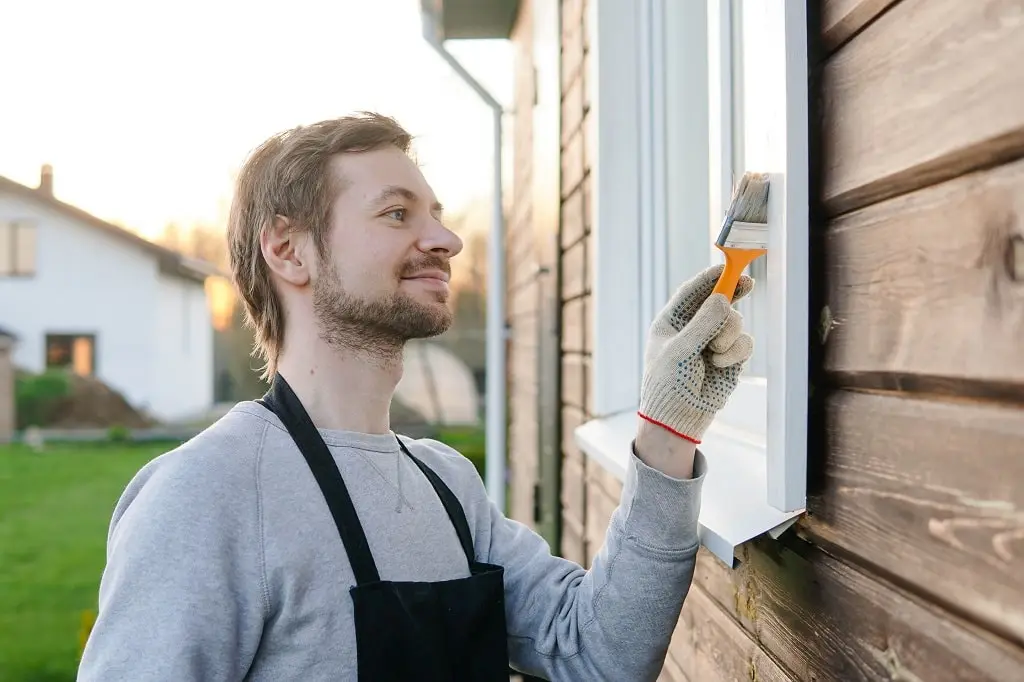 Young smiling man in apron paints the window frame on the exterior of his home.