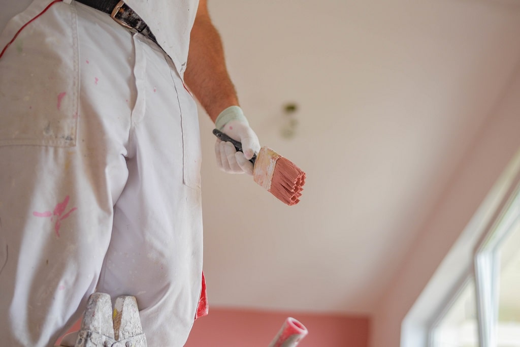The torso of a painter in white pants and tee shirt holding a paint brush with pink paint on it.