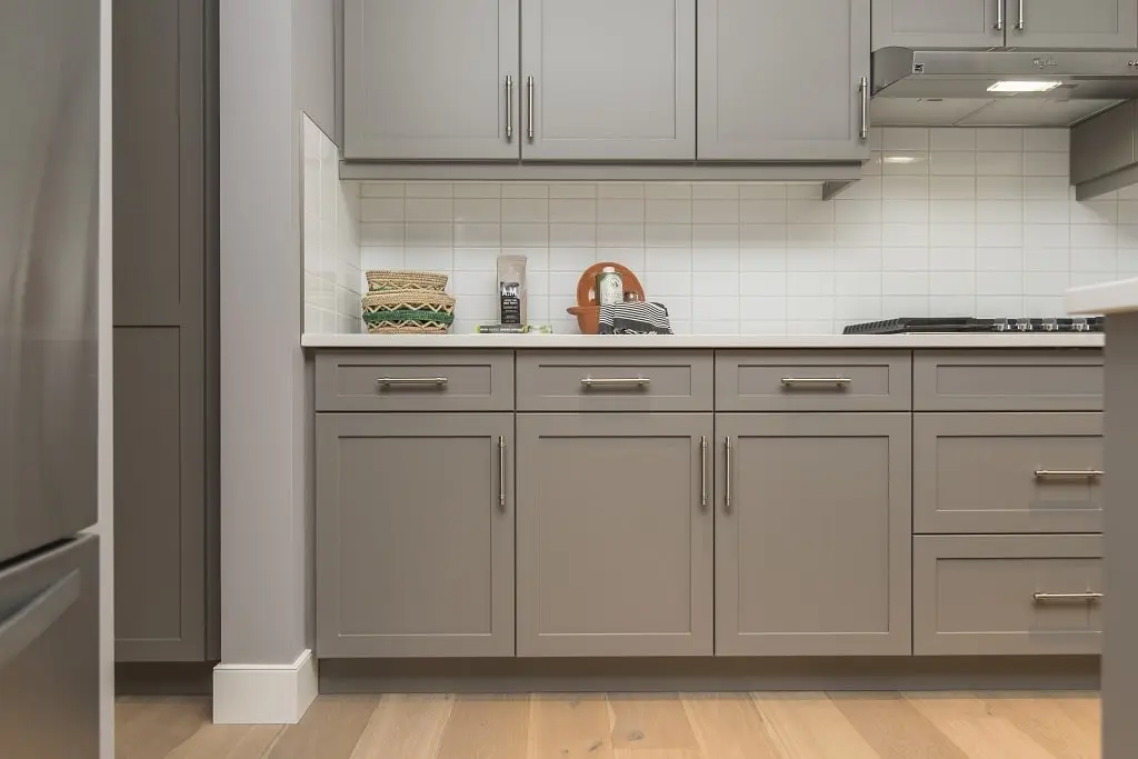 A modern kitchen with gray painted kitchen cabinets, a white tile wall, and wood floors.