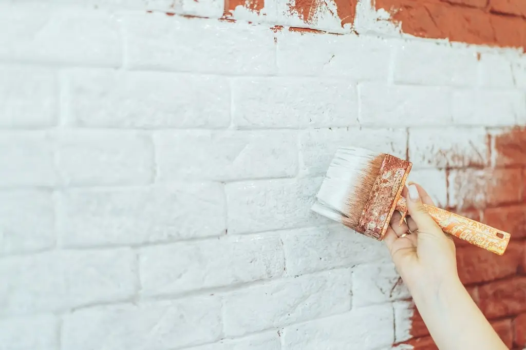 A hand paints an orange brick wall with a paintbrush and white paint.