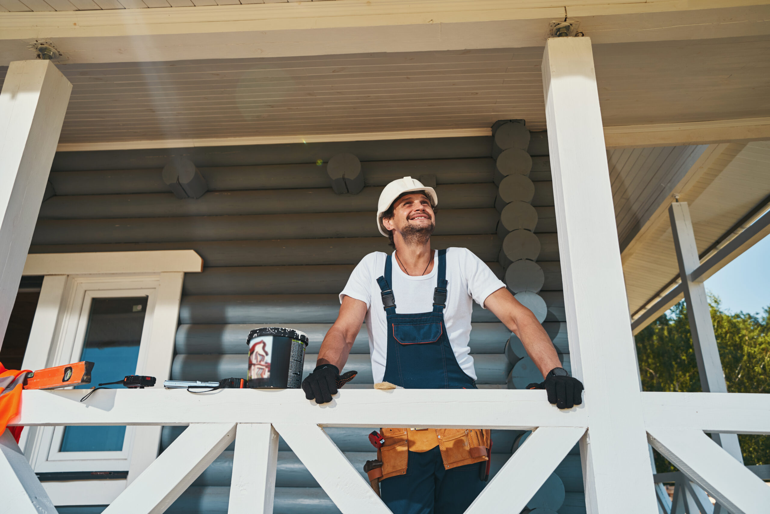 Male in bib-and-brace overalls and helmet looking up at wooden roof of front porch while leaning on fence
